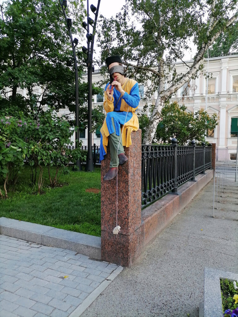 Актер в средневековом костюме шута играет на губной гармошке, сидя на ограждении на бульваре в Москве